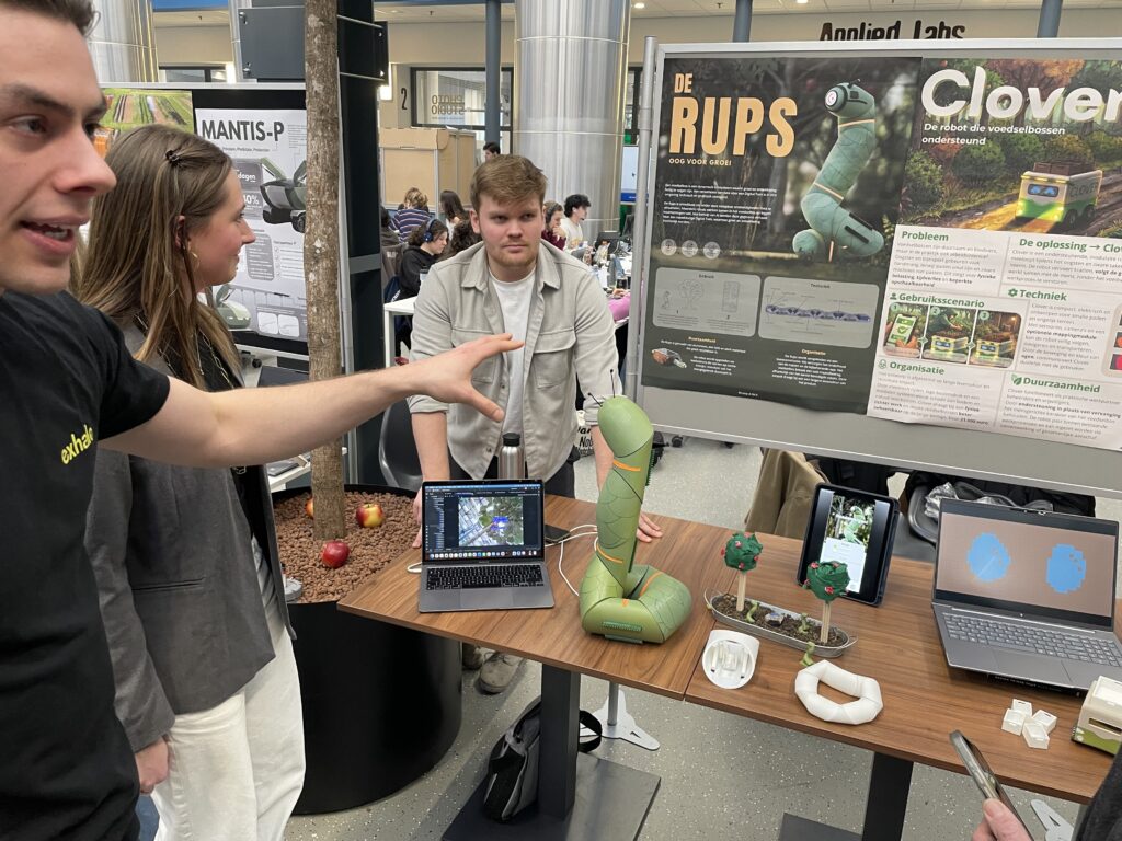 Studenten leggen het concept van de Rups uit, een zwerm van robots die kan rondkruipen door een voedselbos om zo foto's te maken en door middel van (een werkende) AI kan waarschuwen als appels aan de boom rijp zijn.